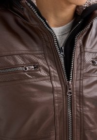 Chaqueta de cuero marrón con cremallera frontal, superficie texturizada con detalles perforados y dos bolsillos en el pecho con cremalleras.
