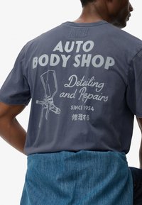 Camiseta de algodón gris con gráfico de "AUTO BODY SHOP" en la parte posterior, que presenta una ilustración de pistola de pintura y texto en japonés. Ajuste y diseño casual.