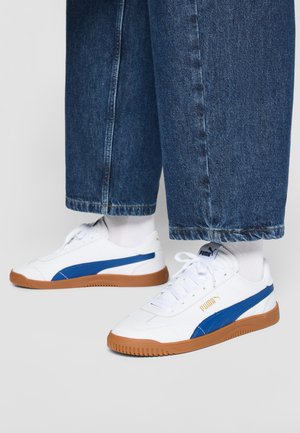 Zapatillas deportivas blancas con acentos azules, suela de goma marrón y superficie texturizada. Combinadas con jeans azul oscuro y calcetines blancos.