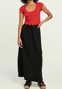 Červený krátký texturovaný top s knoflíky v krátkých rukávech, doplněný černou sukní až na zem s bočními kapsami a šňůrkovým pasem. Černé sandály.