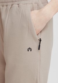 Béžové tepláky so bočnou kapsou a čiernym kovovým doplnkom s logom. Mäkсké, hladké tkaniny s uvoľneným strihom a elastickým pásom.