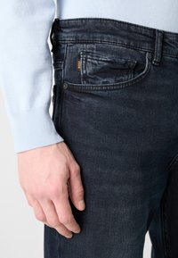 Mano izquierda descansando al lado de unos jeans de mezclilla azul oscuro con una pequeña etiqueta "BOSS" visible cerca del bolsillo, camisa de manga larga azul clara puesta.