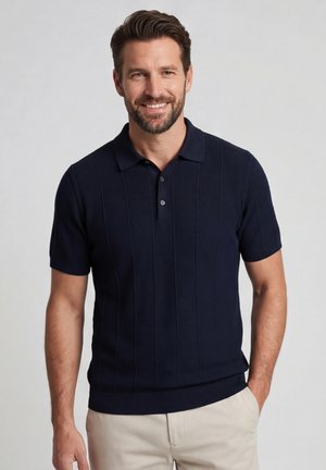 Lachende man met kort bruin haar en baard, draagt een donkerblauw poloshirt met korte mouwen en een beige broek, handen ontspannen langs het lichaam.