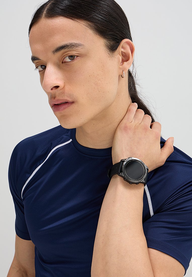 Reloj deportivo digital negro con esfera redonda y bisel texturizado, llevado en la muñeca. Modelo con camiseta atlética azul marino con acentos blancos.