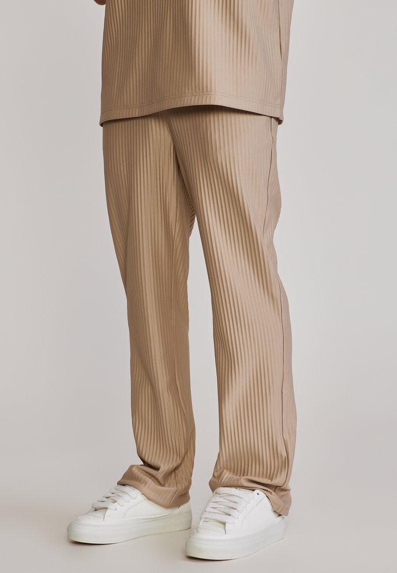 Pantalones beige acanalados y de ajuste holgado combinados con una camiseta a juego y zapatillas blancas bajas sobre un fondo neutro.