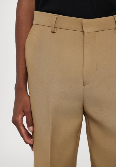 Pantaloni beige su misura realizzati in tessuto liscio, caratterizzati da un frontale piatto, passanti per la cintura e una tasca laterale. Mano appoggiata sulla coscia.