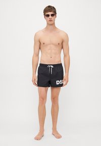 Junger Mann in schwarzen Badehosenshorts mit weißem Kordelzug und "DSQ"-Logo, barfuß vor einem schlichten weißen Hintergrund stehend.
