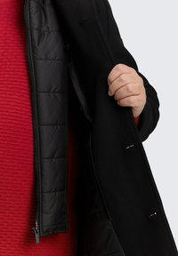 Czarny płaszcz z zamkiem błyskawicznym wewnętrznej warstwy. Wewnętrzna warstwa jest ocieplona pikowanym wzorem. Zewnętrzny materiał wydaje się gładki, z dopasowanym krojem.