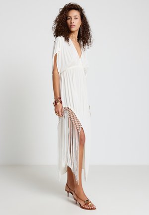 Femme aux cheveux bouclés portant une robe blanche à franges et des sandales à talons marron, debout devant un fond uni.