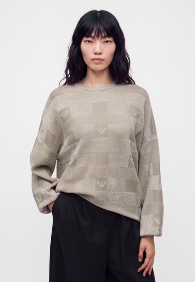 Femme aux longs cheveux noirs portant un pull beige à motifs et un pantalon noir, debout, une main dans la poche, sur un fond uni.