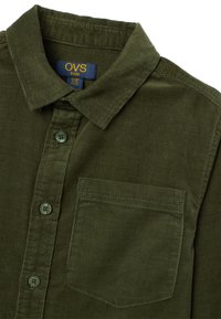 Camisa de pana verde oliva con cuello tipo botón, bolsillo delantero y botones verdes. La textura es acanalada, proporcionando una sensación suave.