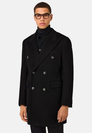 Manteau noir croisé en tissu texturé avec de grands boutons, des revers en pointe et des poches latérales, porté par-dessus un col roulé.