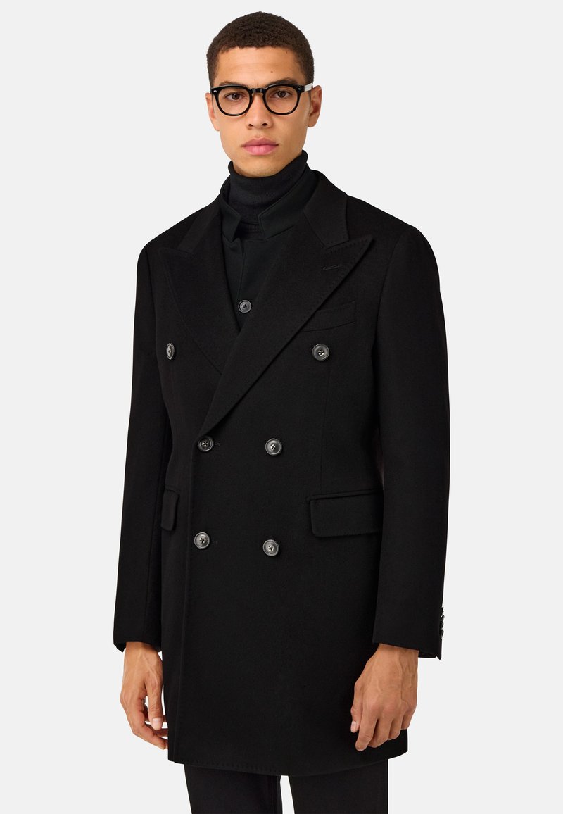 Melna divrindu jaka, izgatavota no teksturēta auduma, ar lieliem pogām, izgrieztām apkaklēm un sānu kabatām, stilizēta virs turtleneck krekla.