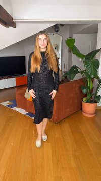 Hosszú szőke hajú nő fekete mintás ruhában és fehér balerinacipőben áll egy fa padlós nappaliban, háttérben nagy zöld növénnyel.