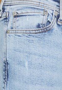 Jeans de ganga azul claro com uma textura desgastada, acessórios prateados e costura em contraste nos bolsos e na cintura.