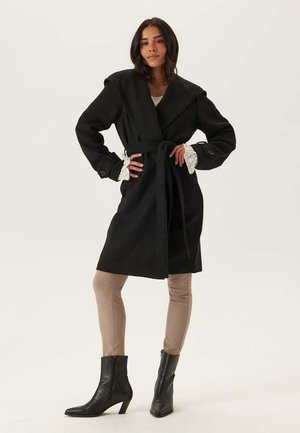 Femme debout avec les mains sur les hanches, portant un manteau noir ceinturé, un pantalon beige, des bottes à talons noires et des poignets en dentelle blanche.