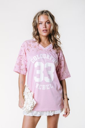 T-shirt rosa in mesh con scritta bianca "33" e "COLOURFUL REBEL", maniche in pizzo e orlo smerlato. Modella che tiene in mano una clutch bianca testurizzata.