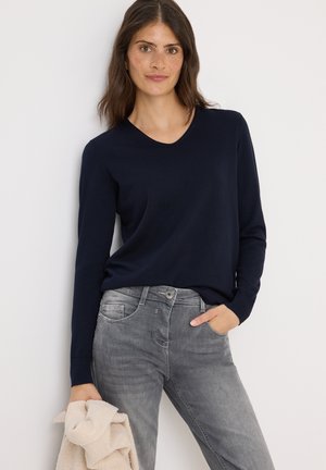 Marineblauwe trui met lange mouwen en een V-hals, gecombineerd met lichtgrijze jeans. De trui heeft een soepele textuur en aansluitende mouwen.