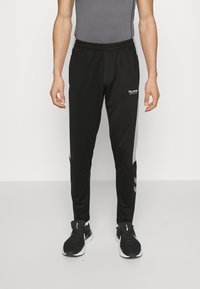 Schwarze Sporthose mit elastischem Bund, ausgestattet mit reflektierenden Seitenelementen und schmalem Schnitt. Getragen mit schwarzen Laufschuhen.
