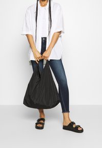 Bolso tote de nylon negro sostenido en la mano, con un diseño suave y flexible. La modelo lleva una camisa blanca holgada y unos jeans ajustados azules con sandalias negras.