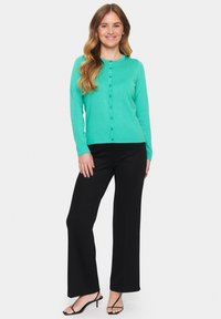 Mündroheline kardigan ümmarguse kaelusega, esipainutite ja pikkade varrukatega, mis on kombineeritud mustade laiadest pükstest ja mustadest sandaalidest.