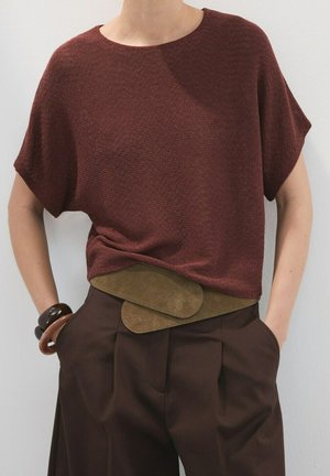 Persona con top de punto marrón de manga corta, pantalones plisados de tiro alto en marrón oscuro, cinturón ancho de ante verde oliva y pulseras gruesas, con las manos en los bolsillos.