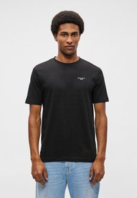 Melns kokvilnas T-krekls ar klasisku apkakli, īsām piedurknēm un mazu baltu logo kreisajā krūtīs; kombinēts ar gaiši ziliem džinsiem.