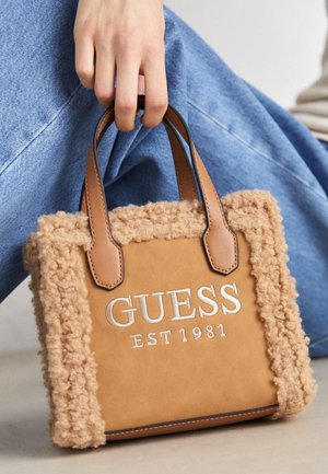 Hand holding a tan faux shearling handbag with leather handles and white embroidered "GUESS EST 1981" text, next to person in blue jeans.