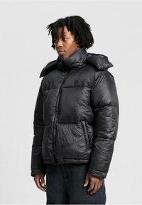 Mand med medium-mørk hud iført en sort vinterjakke med høj krave og lynlås, stående foran en ensfarvet lysegrå baggrund.