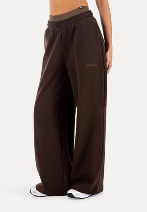 Person trägt dunkelbraune, weit geschnittene Jogginghosen mit elastischem Bund und weiße Turnschuhe, steht vor einem einfarbigen Hintergrund.