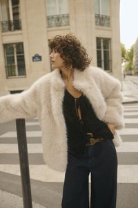 Šviesiai smėlinės spalvos dirbtinio kailio paltas su aukštu apykakle, dėvimas ant juodos bliuskės su volanais. Sujungtas su tamsiai mėlynomis plačiomis džinsomis.