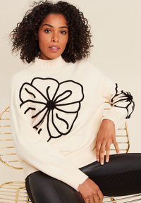 Maglione color crema con un grande motivo floreale nero sulla parte frontale e sulle maniche. Presenta un collo alto e polsini a coste.
