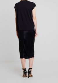 Frau in einem schwarzen ärmellosen Top, einem schwarzen Midirock mit einem Schlitz auf der Rückseite und schwarzen High-Heels mit Knöchelriemen, die vor einem schlichten Hintergrund steht.