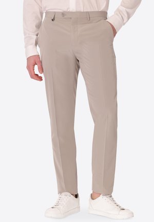 Uomo che indossa pantaloni beige su misura, camicia bianca e sneaker bianche, con una mano in tasca e un braccio rilassato.