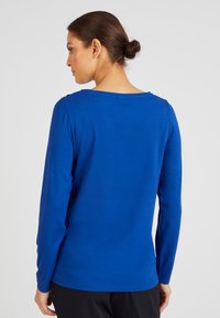 Camicetta a maniche lunghe in tessuto blu brillante. Collo a girocollo con dettagli a bottone sulle spalle. Texture morbida e design aderente. Colore semplice e uniforme.