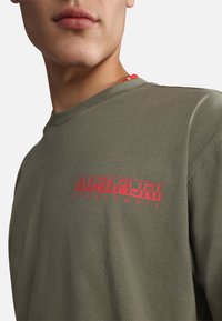 Camiseta de algodón verde oliva con un logo de texto rojo en la parte delantera. El diseño incluye un cuello redondo y mangas cortas.