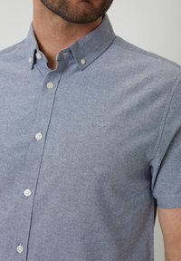 Camisa de manga corta, con botones, en tejido azul claro texturizado. Presenta botones blancos y un pequeño logo bordado en el pecho.