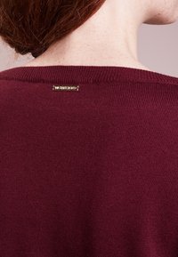 Maglione a maglia bordeaux con colletto a coste, dotato di un piccolo logo in metallo dorato sul retro e una finitura a trama liscia.