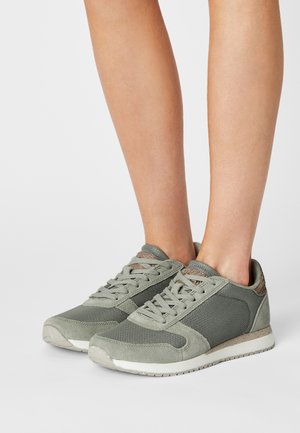 Feet wearing gray and beige mesh and suede sneakers laced up, standing against a plain white background.