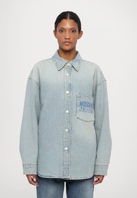 Gaiši zils džinsu krekls ar pogām priekšpusē, norādītu apkakli un krūšpockets, uz kura ir uzraksts "MOSCHINO JEANS" zilos burtiem. Brīvs piegriezums.