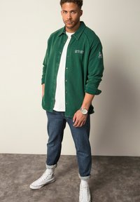 Grøn skjorte med hvid broderi, lagd over en hvid t-shirt, parret med rullede jeans og hvide højhælede sneakers.