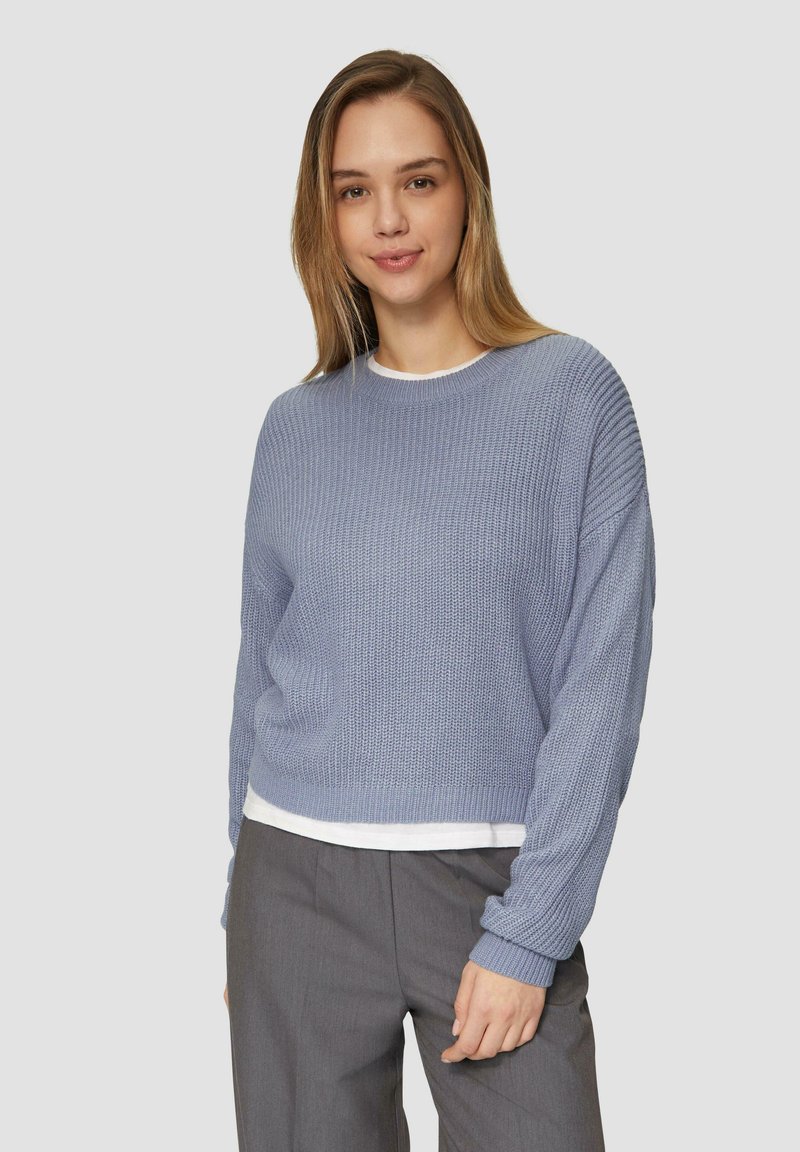 Hellblauer, gestrickter Pullover mit strukturiertem Rippenmuster, rundem Halsausschnitt und langen Ärmeln. Über einem weißen Hemd getragen und mit grauen Hosen kombiniert.