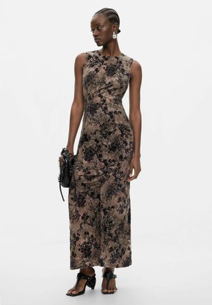 Femeie în rochie fără mâneci, cu imprimeu floral, până la gleznă, ținând o geantă neagră, purtând sandale negre cu toc și cercei argintii, pe fundal alb.