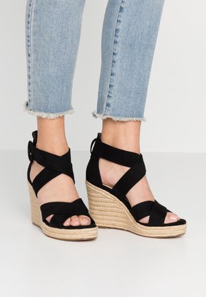 Czarne sandały na koturnie z plecionym korkiem. Posiadają krzyżujące się paski oraz zapięcie na kostce, podkreślone miękką teksturą z zamszu.