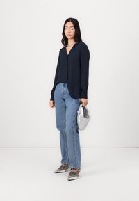 Marineblå langermet bluse med avslappet passform, kombinert med høyt liv lyseblå jeans, sølvmetalliske sko og en sølvfarget knyttet håndveske.