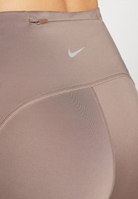 Leggings atléticos marrones hechos de material elástico, con una textura suave y un pequeño logo gris de Nike en la cinturilla.