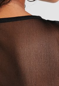 Top de malla negra con un tejido texturizado y semitransparente; presenta un escote negro sólido y un corte holgado.