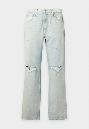 Jeune homme debout, une main dans la poche, portant une chemise beige à manches courtes boutonnée, un jean clair déchiré et des chaussures marron décontractées, sur fond blanc.