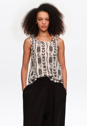 Kobieta o kręconych włosach, ubrana w bezrękawnik w wzory i luźne czarne spodnie, stoi z rękami w kieszeniach na białym tle.