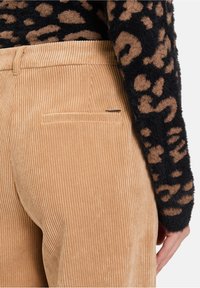 Tijgerkleurige corduroybroek met verticale ribbels, voorzien van een enkele achterzak en een slanke metalen accent. De stof heeft een zachte textuur.